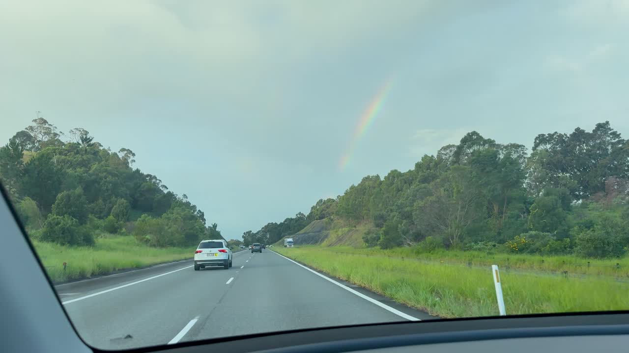 A car travels along a lush, green highway under overcast skies, approaching a faint rainbow on the horizon. Natural light, steady dashboard perspective
