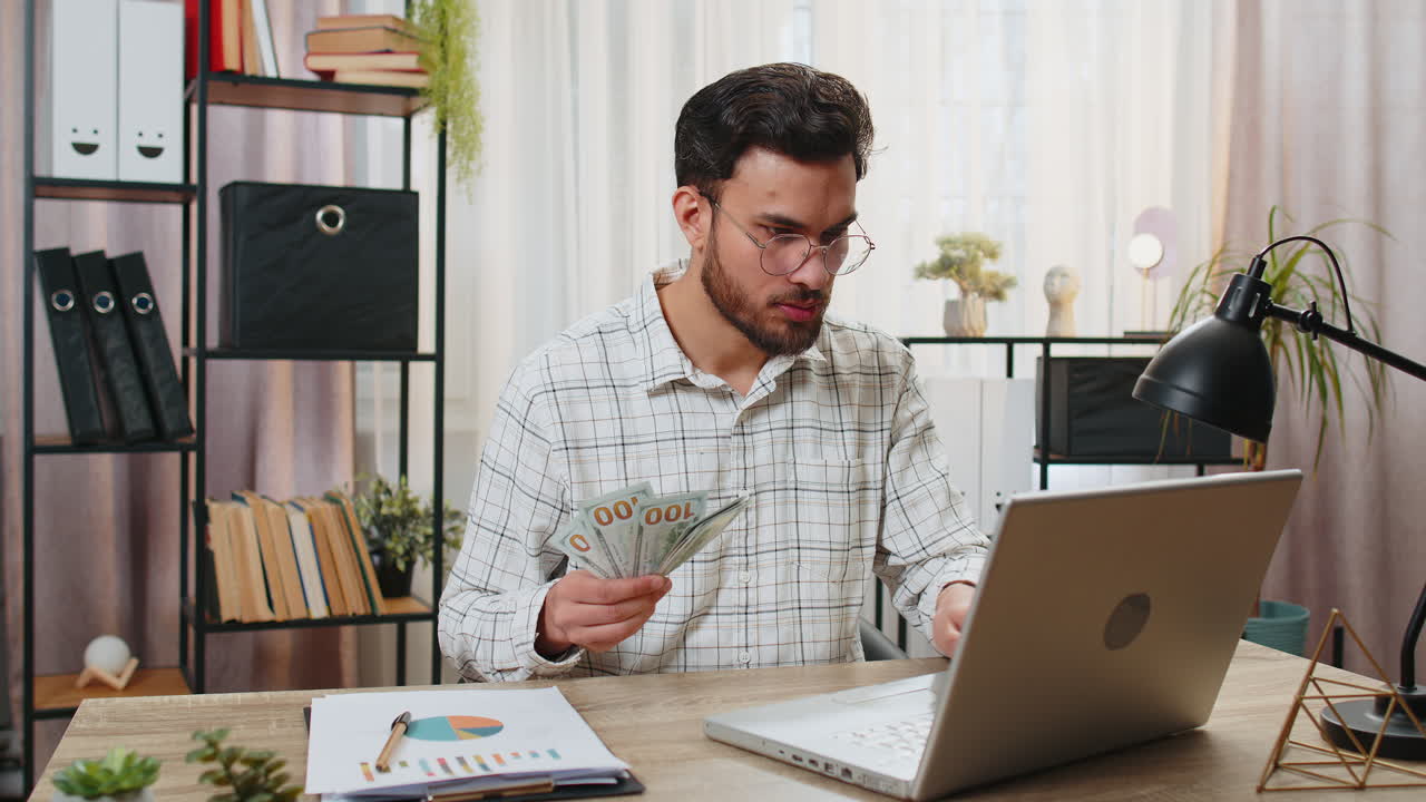 Hombre de negocios indio contando dinero en efectivo calcula dividendos y usando portátil en la oficina en casa