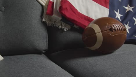 Lounge-Zu-Hause-Einer-Person,-Die-Ein-American-Football-Spiel-Im-Fernsehen-Mit-Stars-And-Stripes-Flagge-Und-Ball-Anschaut-2