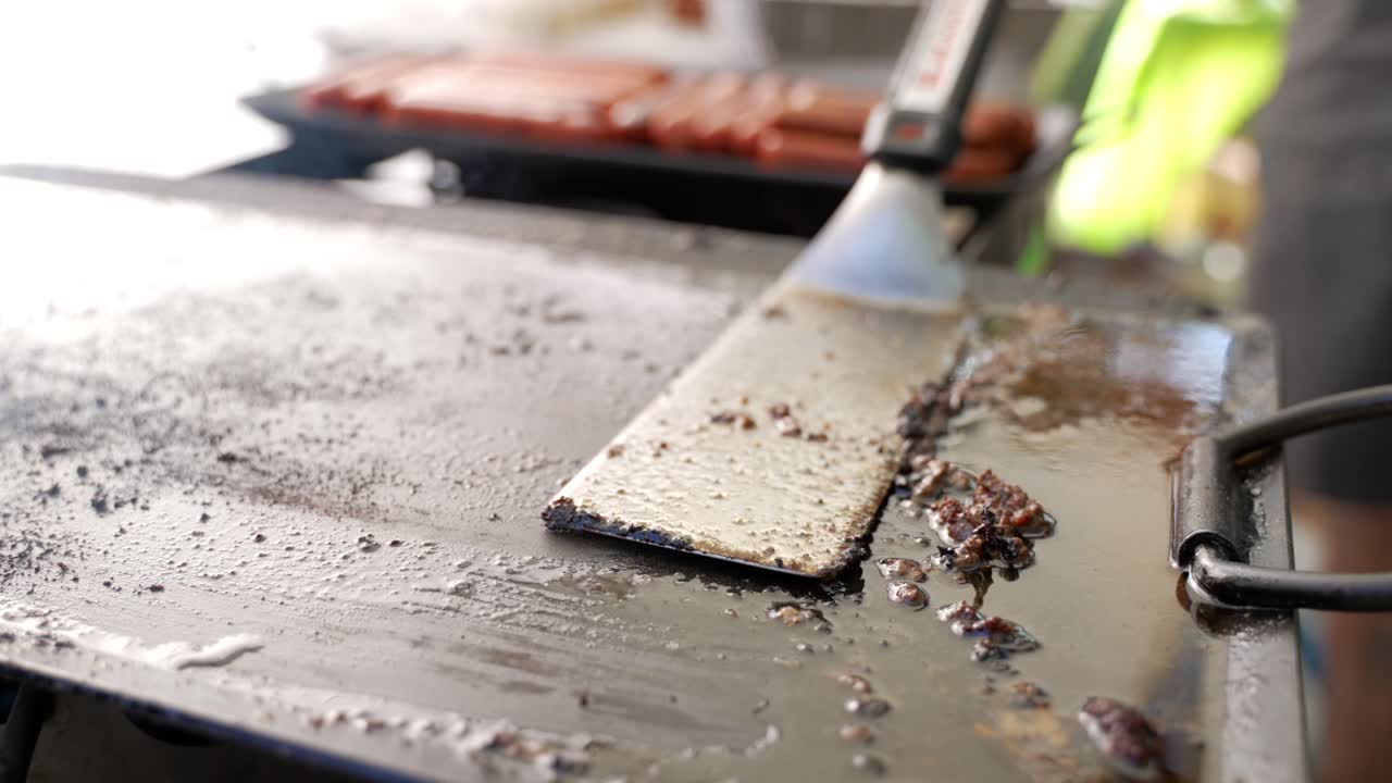 Setting spatula down on greasy meat covered cook top grill with hot dogs sausages and veggie wieners in background at outdoor campsite barbecue