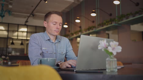 A-young-man-in-a-shirt-is-sitting-at-a-table-with-a-laptop-and-typing-on-the-keyboard.-A-student-can-study-remotely.-A-businessman-conducts-his-business-remotely