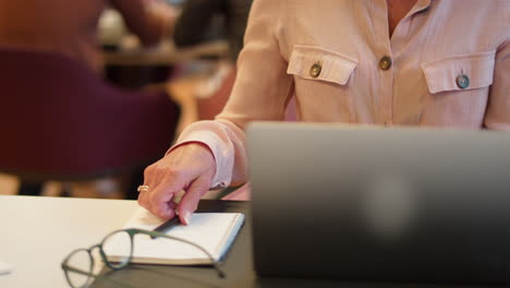 mature businesswoman working on laptop at desk in office making notes in notebook