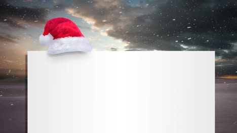 Animation-of-blank-card-with-copy-space-and-santa-hat-over-snow-falling-and-winter-scenery