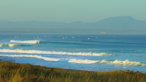 Cinematic-pan-stunning-early-morning-huge-waves-glassy-swell-surf-Hossegor-Seignosse-France-yellow-sunrise-sunset-on-beach-mountain-coast-Biarritz-Basque-Country-grassy-sandy-shorline-sailboats-at-bay