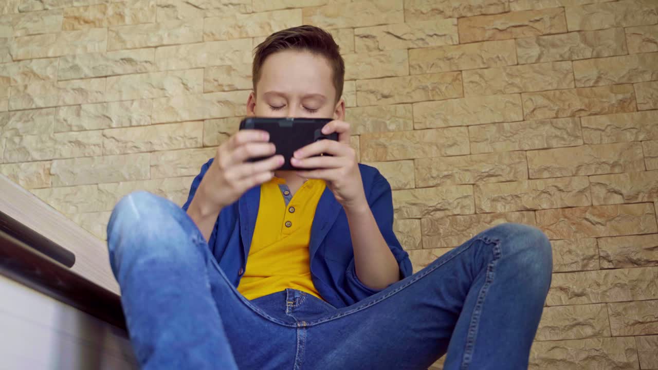 Portrait of a boy with a smartphone at home. Teenager is playing on his phone while sitting on stairs. Social distancing and self isolation in quarantine.