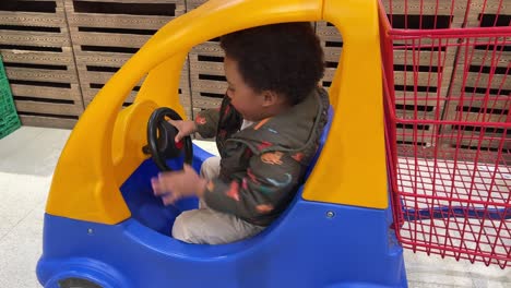 playful two year old back baby rides in a blue and lyelllow shopping trolley inside a supermarket