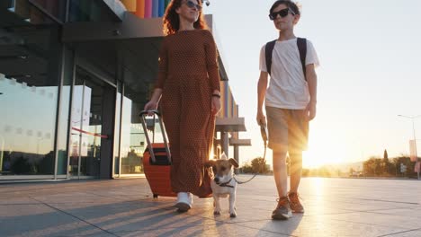 happy boy, woman with an orange suitcase in long dress and dog jack russell walking along terminal of airport in lens flares sunset in summer in slow motion. family goes on trip. travel. tourism