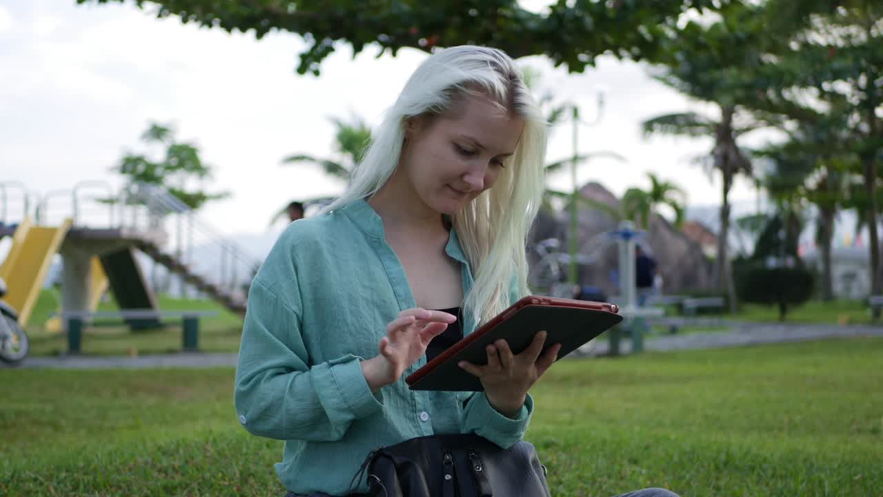 hermosa mujer delgada con el cabello largo rubio en camisa verde se sienta en el suelo y el uso de teléfono inteligente sobre el fondo del parque. niña en la pantalla cuadrada tocando y sonriendo