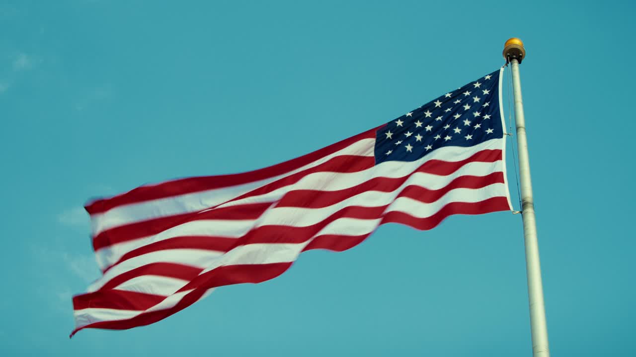 USAs flagg som vaier på en flaggstang mot blå himmel, patriotisk symbol på frihet og demokrati.