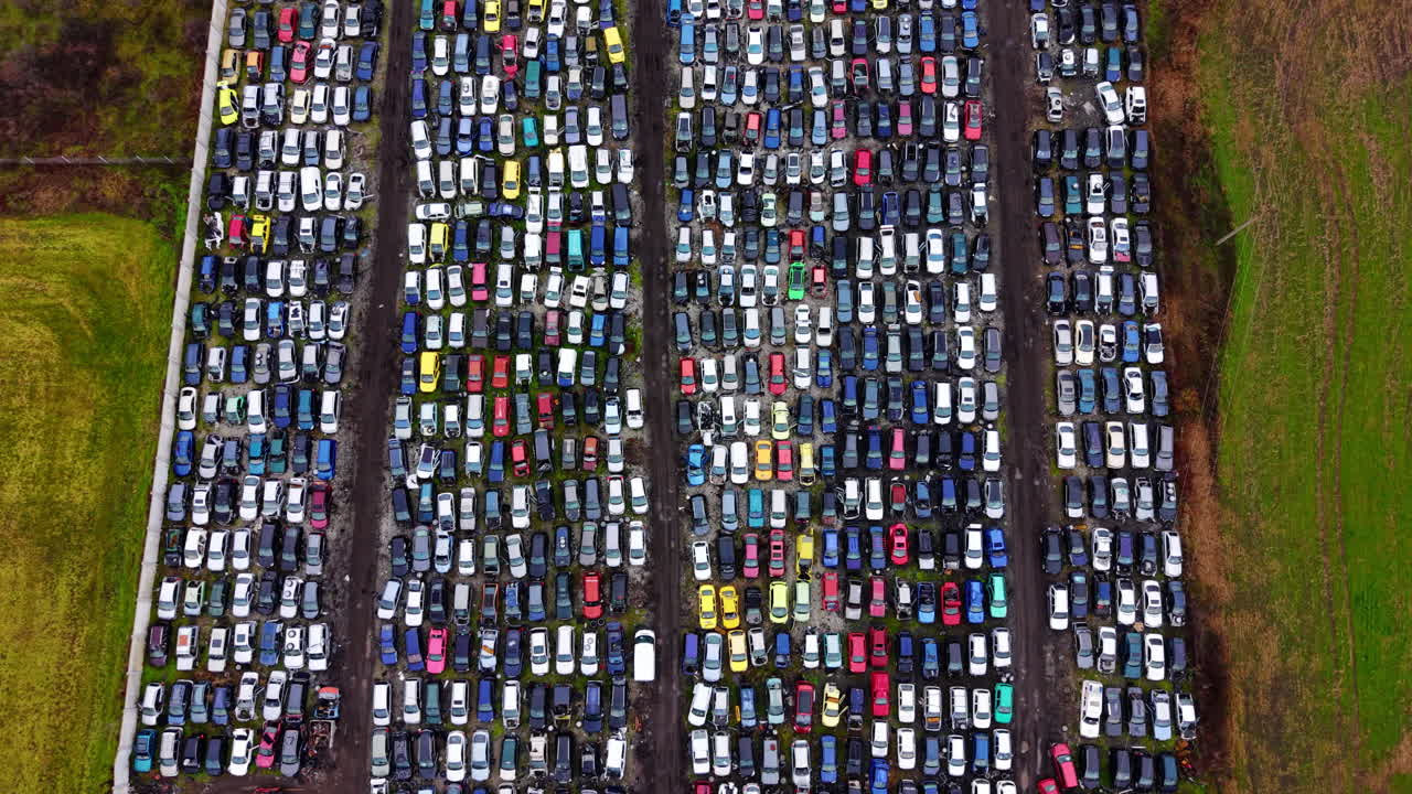 Toma aérea de un gran desguace de coches con vehículos clasificados en filas separados por caminos de tierra, destacando la organización industrial. Vista superior simétrica de coches en una instalación de reciclaje