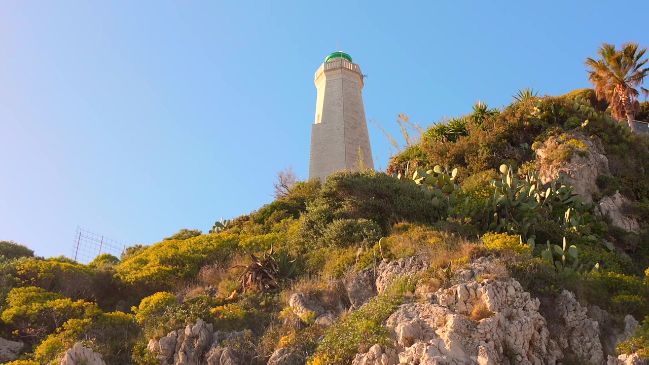 Faro di Cap-Ferrat che si erge sopra aspre scogliere costiere sotto un cielo azzurro limpido, accanto all'ambiente mediterraneo a Saint-Jean-Cap-Ferrat, Alpi Marittime, Francia.