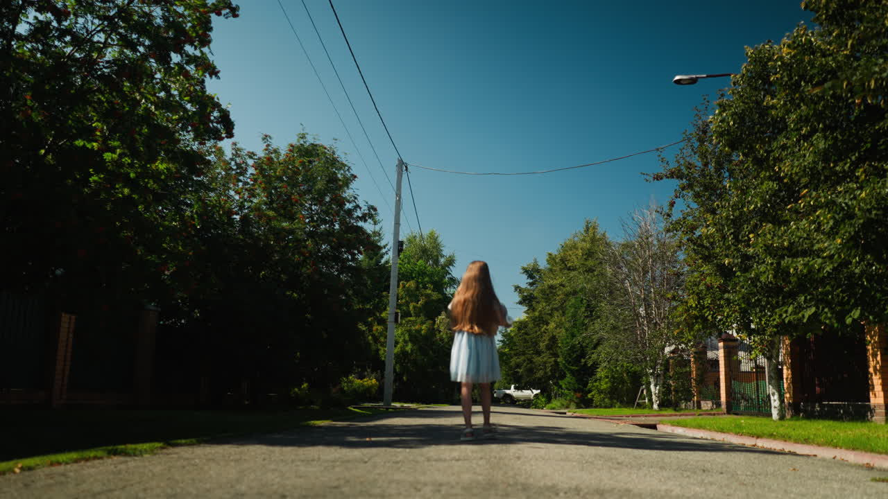 Rear view of girl with long hair walking calmly down quiet residential street on sunny day, holding toy in hand as butterfly passes by, surrounded by trees, fences, and distant parked car