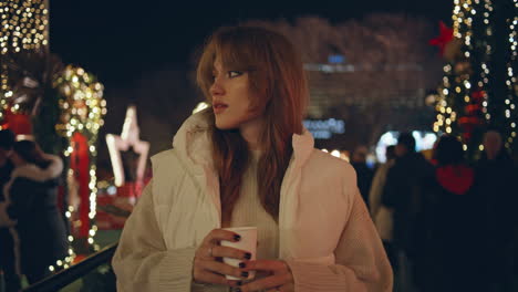 mujer caminando en la feria de navidad sosteniendo una bebida caliente por la noche luces festivas de primer plano.