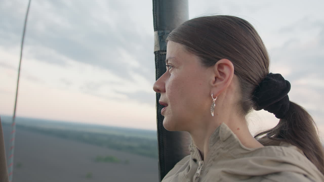 woman holds radio handset while looking down over sprawling farmland from hot air balloon basket at dusk haze capturing communication and aerial perspective with pink sky and open field view