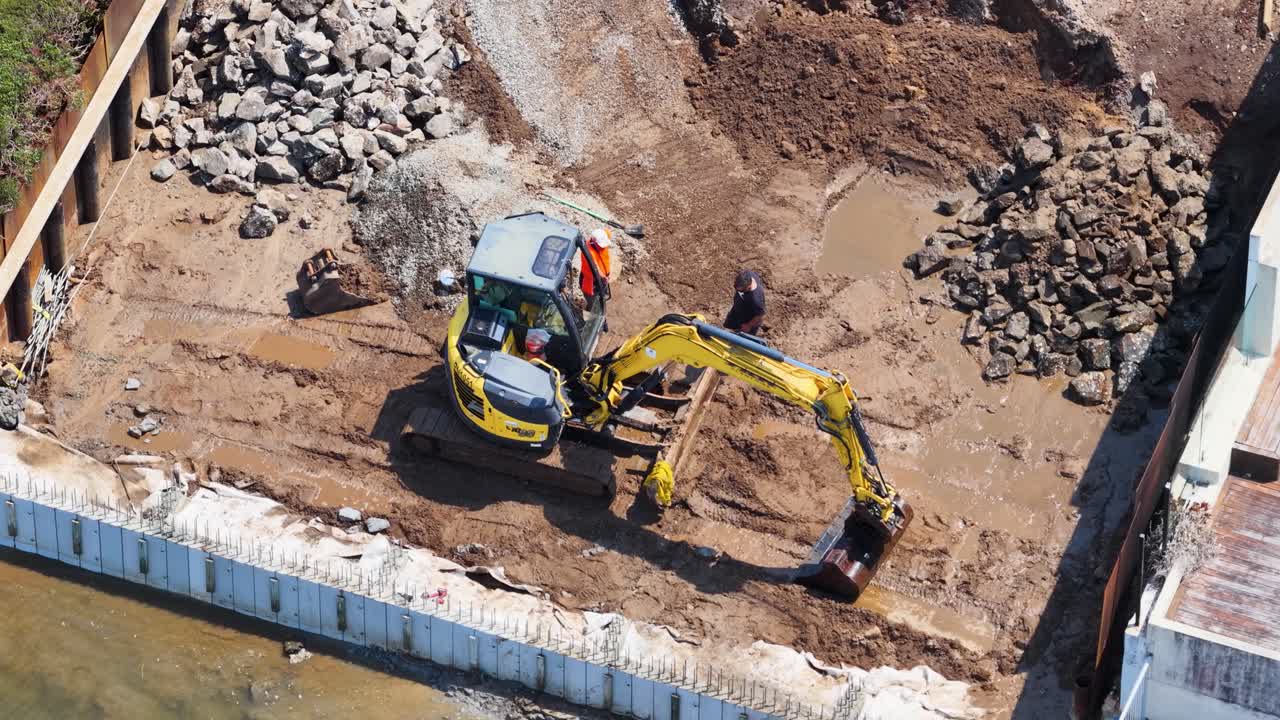 Drone shot of mini excavator and workers digging muddy waterfront foundation in bright daylight
