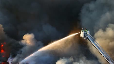 Filmische-Drohnenaufnahme-Von-Zwei-Feuerwehrleuten-Auf-Einer-Leiter,-Die-Wasser-Auf-Ein-Brennendes-Gebäude-Sprühen,-Während-Dunkle,-Giftige-Dämpfe-Aufsteigen-–-Zoomaufnahme-Von-Oben-Nach-Unten-Aus-Der-Luft