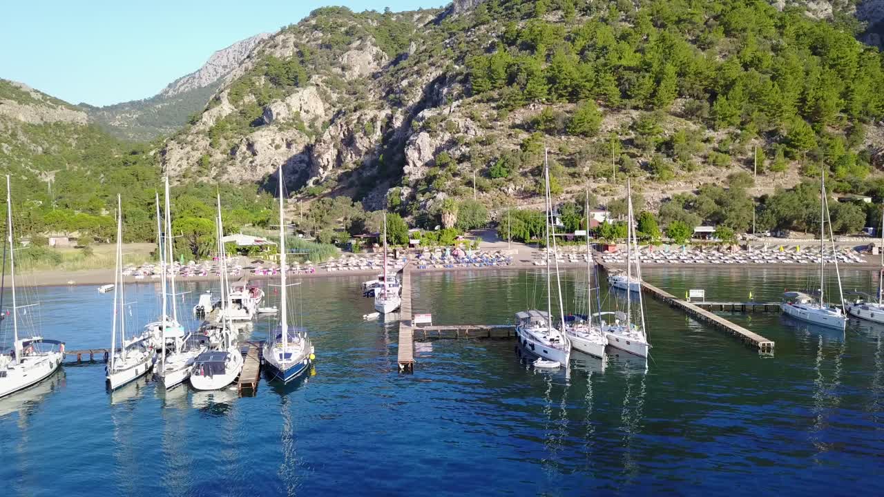 Aerial view of Cifltik bay,Footage forward towards the beach full with sunbeds and green hills.Sailboats stern-to restaurant's piers on calm sea.