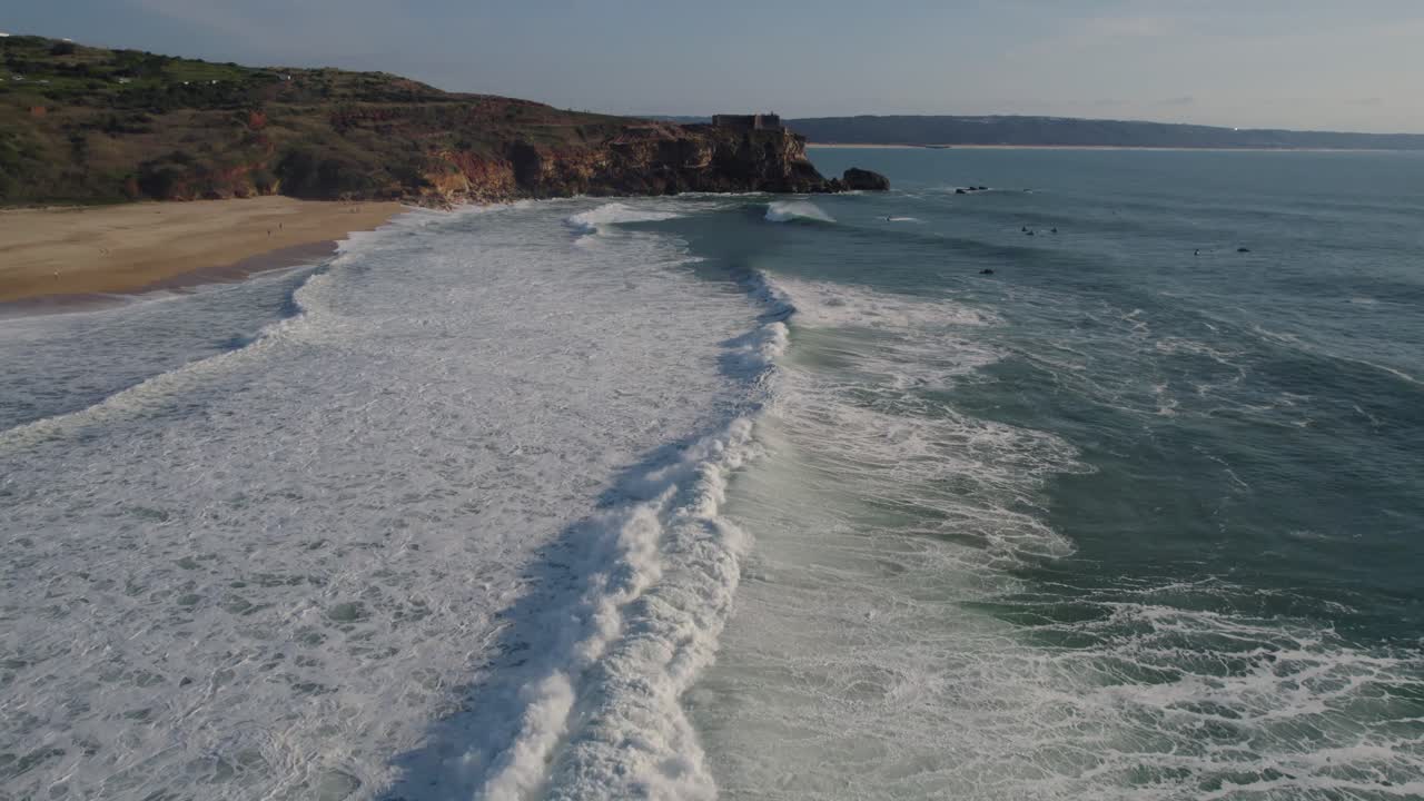 las olas chocando contra la playa de arena de la playa de nazare con altos acantilados rocosos que bordean la costa en el fondo