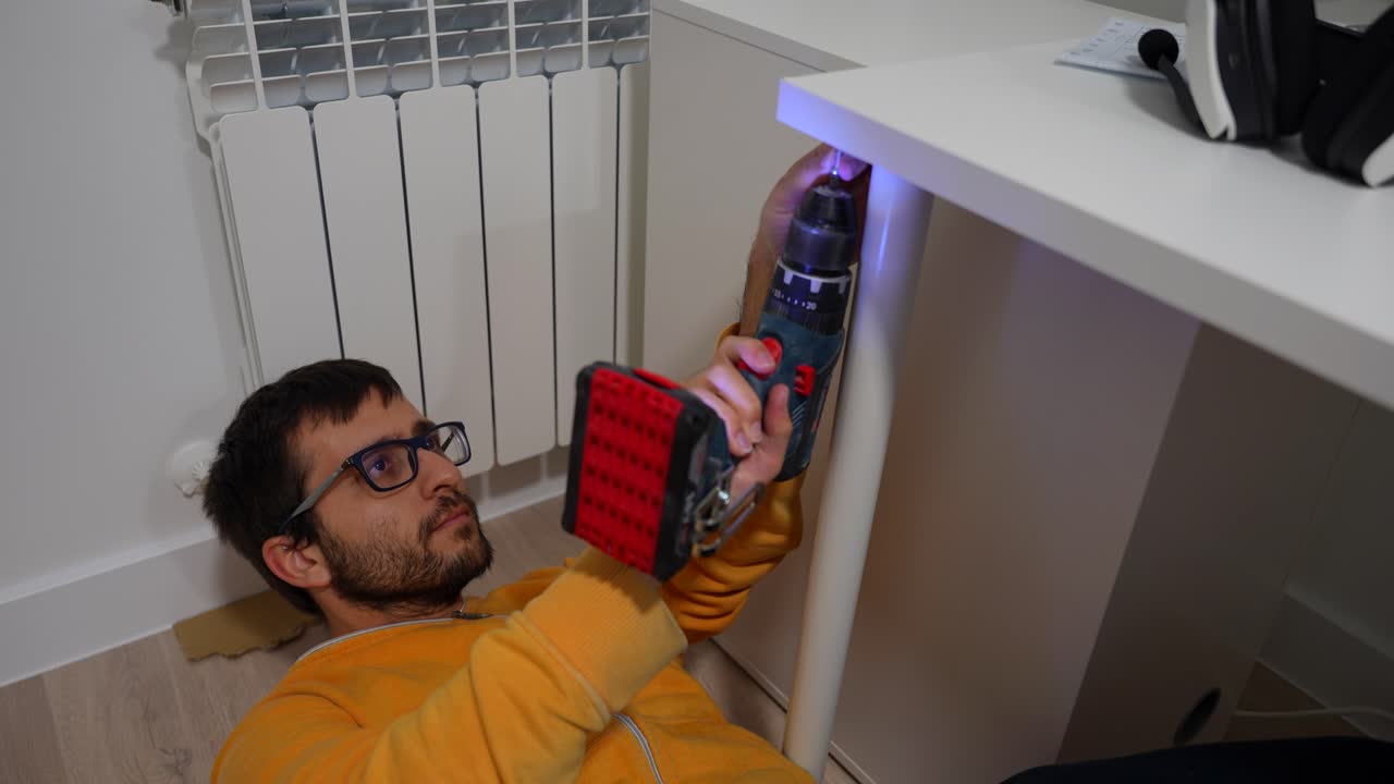 Focused young man lying on the floor fastening a leg to table with electric screwdriver