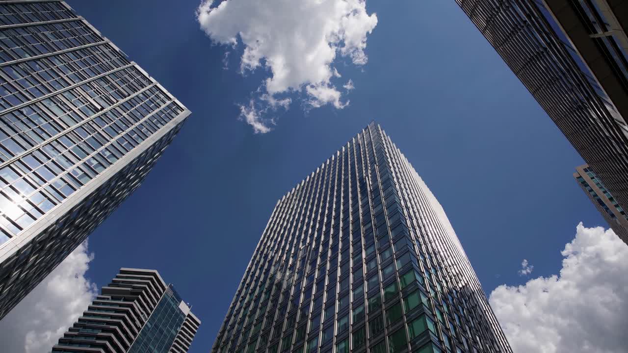Filmagem em vídeo de ângulo baixo de arranha-céus modernos contra um céu azul com nuvens, enfatizando o cenário urbano.