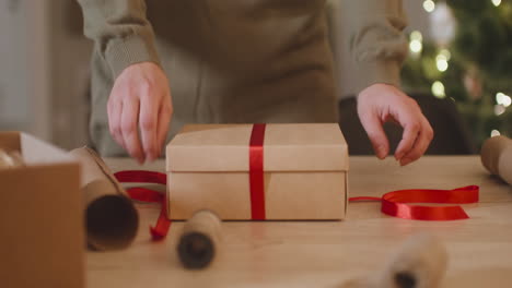 die kamera konzentriert sich auf die hand einer frau, die weihnachtsgeschenke auf einem tisch in einem mit einem weihnachtsbaum geschmückten raum einwickelt 2