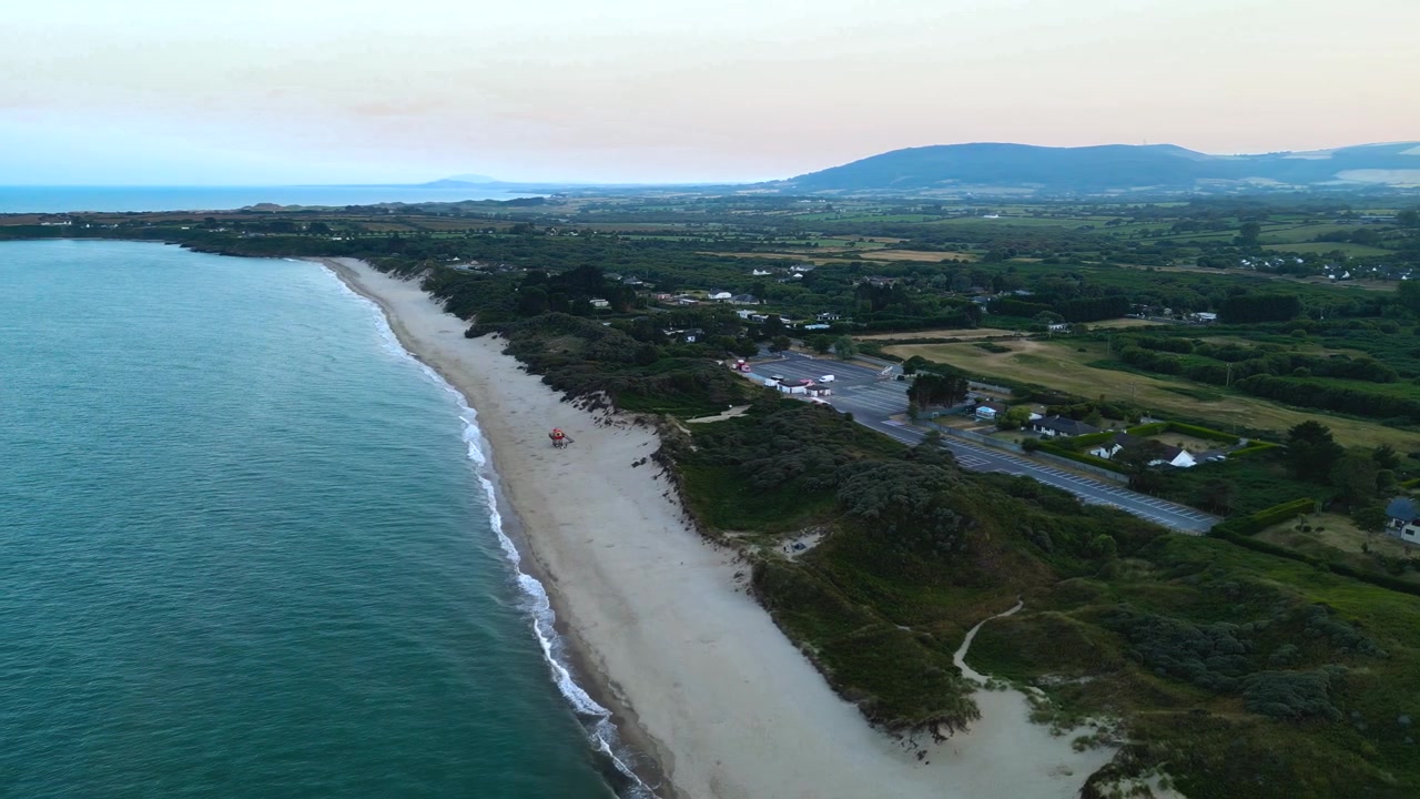 Drone che effettua una panoramica a sinistra su Brittas Bay, rivelando una spiaggia sabbiosa, onde leggere, dune, campi verdi e uno sfondo montuoso al tramonto