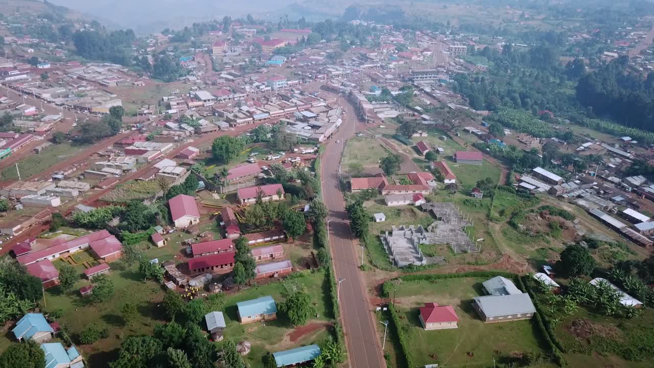Drone shot over Kapchorwa town shows main paved road, rooftops, guesthouses, and administrative buildings nestled in the hilly terrain of Mount Elgon, with visible banana groves and farmlands