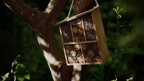 In-the-midst-of-a-lush-green-garden,-a-captivating-wooden-beehive-hangs-from-a-tree,-housing-industrious-bees-amidst-the-summer-scenery