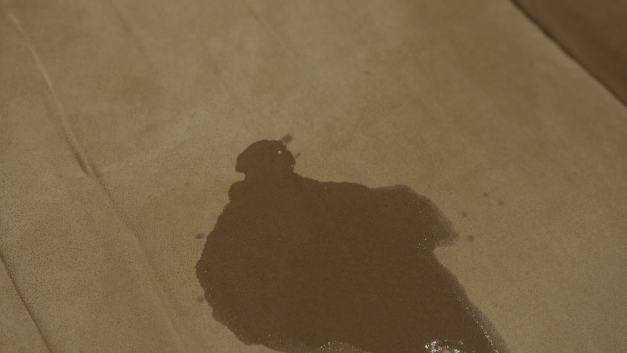 A close-up of a wet stain on a brown fabric sofa. The fabric darkens as the liquid spreads, showing the effect of moisture on upholstery