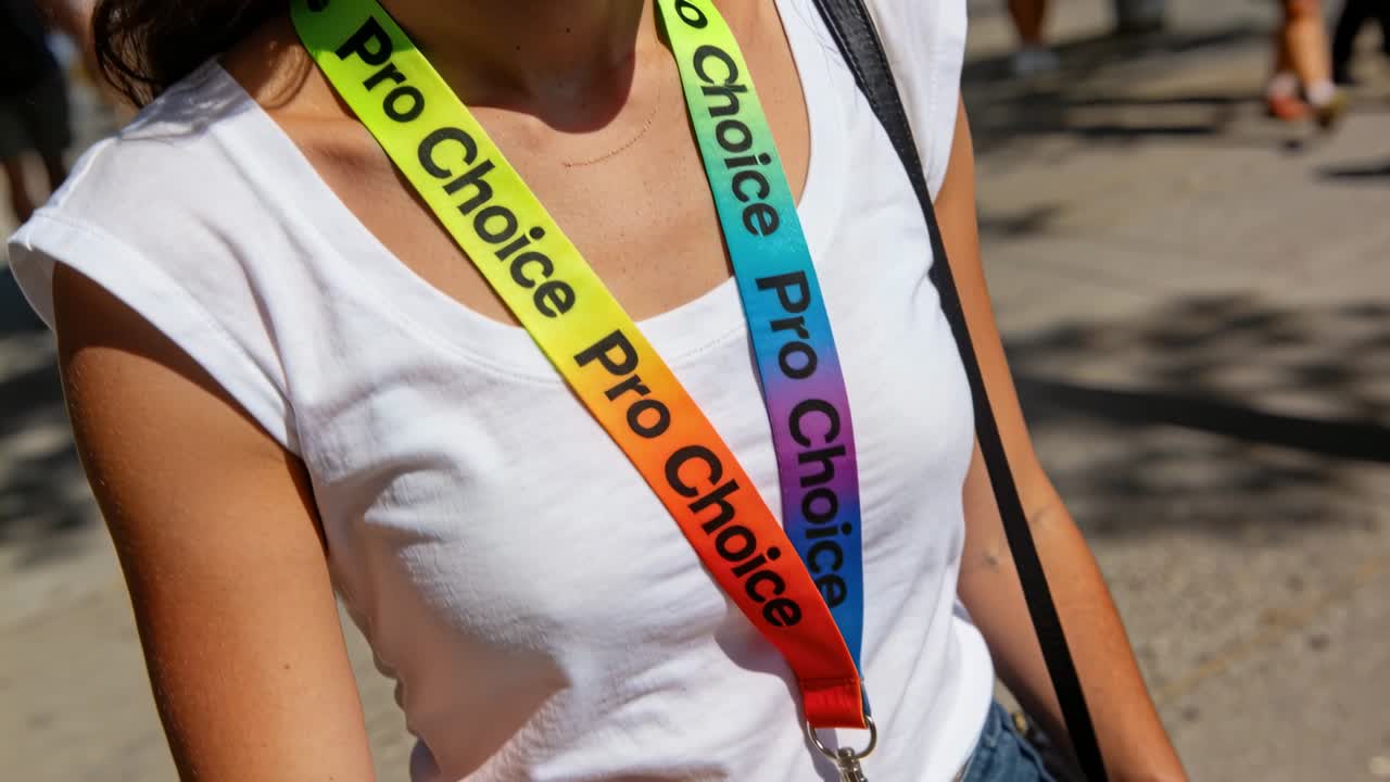 Gehende Frau in weißem T-Shirt auf sonnigem Weg, Kamera bewegt sich näher und zeigt einen Regenbogen-ProChoice-Schlüsselband-Ausweis.