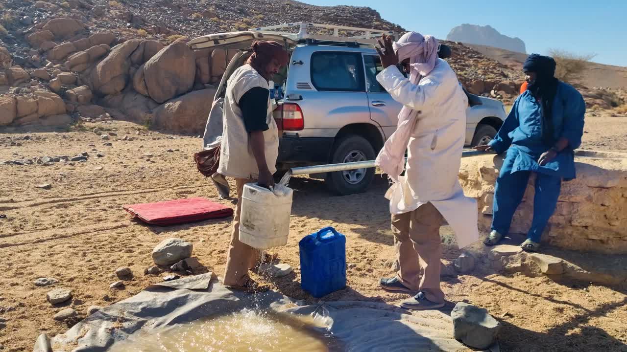 Local Tuareg man filling water container with drinking water, meanwhile talking, smoking and having fun in Sahara desert on windy sunny day in Tamanrasset region of Algeria