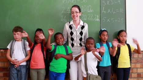 Pupils-and-teacher-waving-and-smiling-at-camera-in-classroom