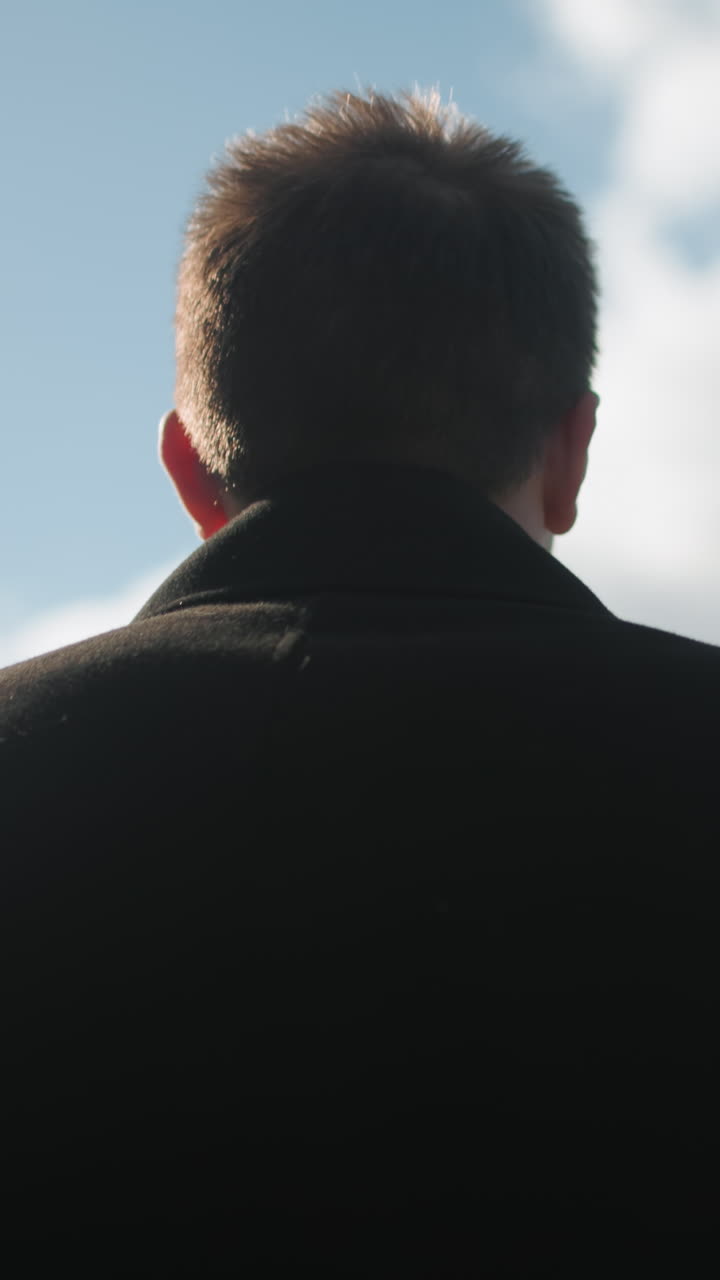 현대 도시 건축과 초록색으로 둘러싸인 구름이 있는 밝은 파란 하늘 아래에서 외부에서 자신감 있게 고 있는 검은 코트를 입은 남자의 뒷면