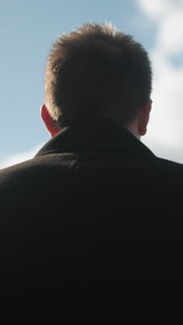 back view of man in black coat walking confidently outdoors under bright blue sky with clouds, surrounded by modern urban architecture and greenery