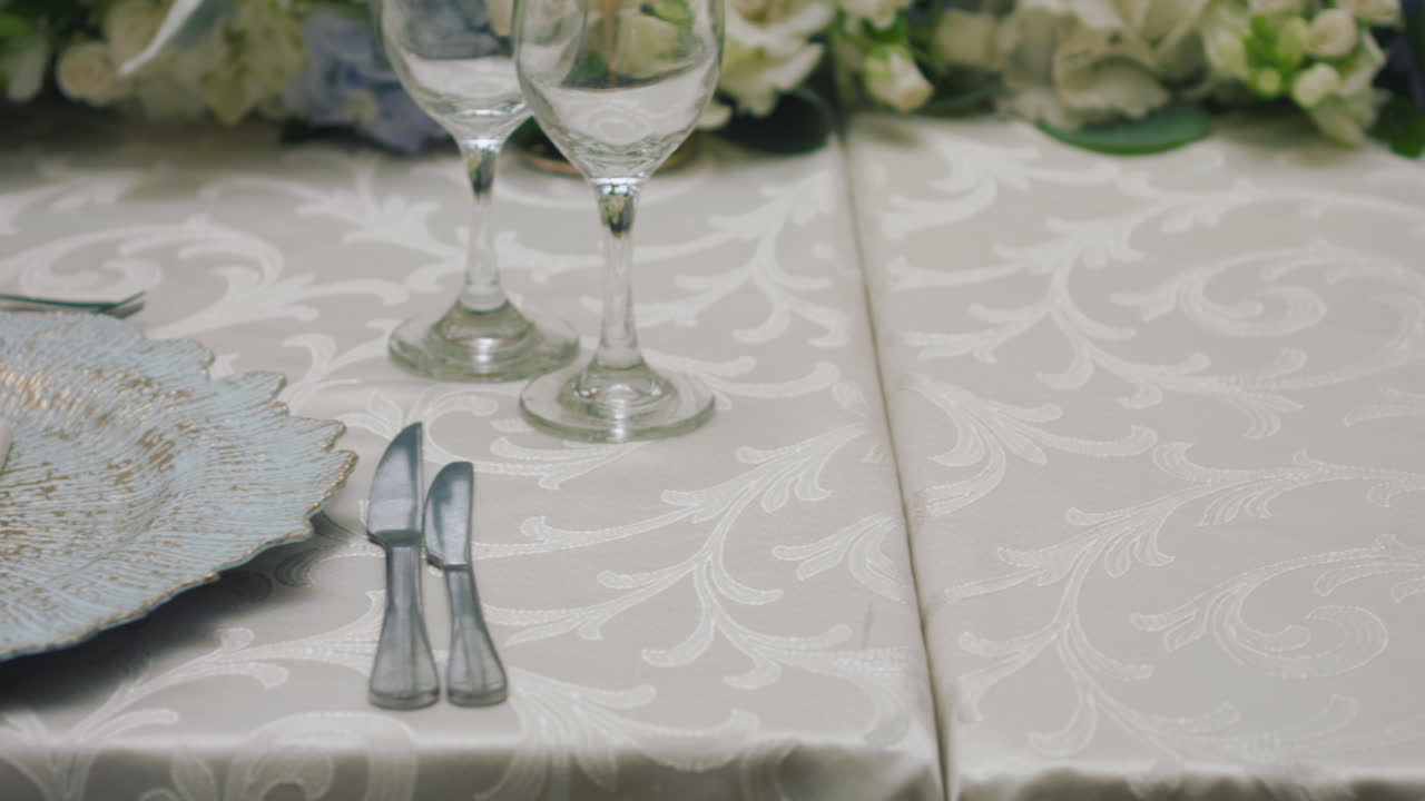 A detailed clip of an elegantly decorated wedding table. It features stemware, a white tablecloth, plates, cutlery, cloth napkins, a delicate floral arrangement, and a personalized invitation.