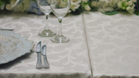 a detailed clip of an elegantly decorated wedding table. it features stemware, a white tablecloth, plates, cutlery, cloth napkins, a delicate floral arrangement, and a personalized invitation.