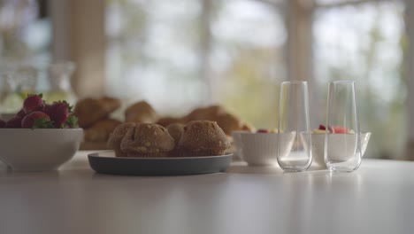 a woman's hand puts down a plate of muffins on a kitchen counter with other breakfast food in the background - in slow motion