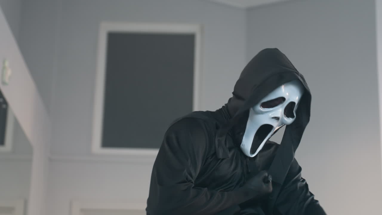 masked figure in black robe theatrically plunges knife into chest then collapses in slow motion on studio floor with mirror reflection and stools in background