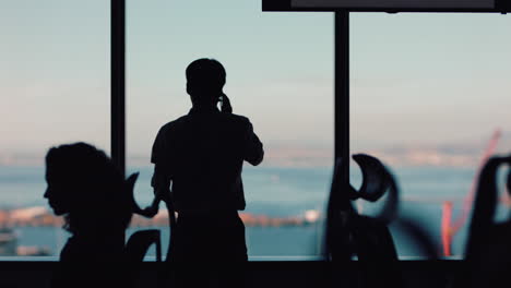 silueta de un hombre de negocios que usa un teléfono inteligente haciendo una llamada telefónica hablando por teléfono móvil mirando por la ventana un ejecutivo corporativo discutiendo ventas trabajando hasta tarde en una oficina ocupada