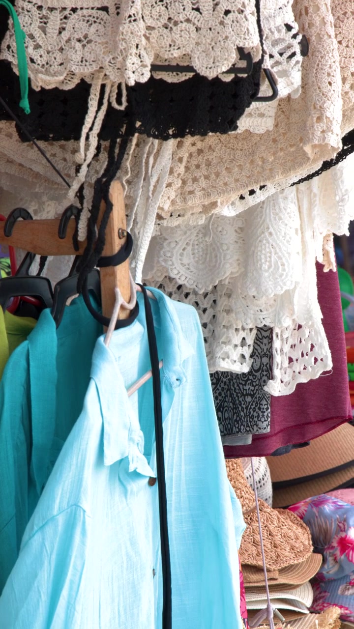 Ripresa panoramica ravvicinata di camicie vivaci, bikini e capi in pizzo appesi a un mercato.