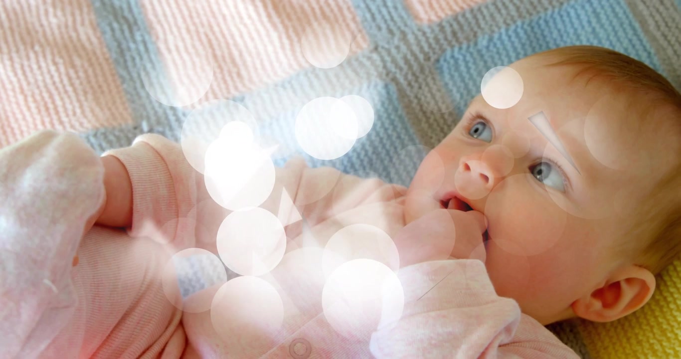 Jentebaby som ligger og reagerer på drivende bokeh-former ved å følge med og sutte på fingrene for god helse.
