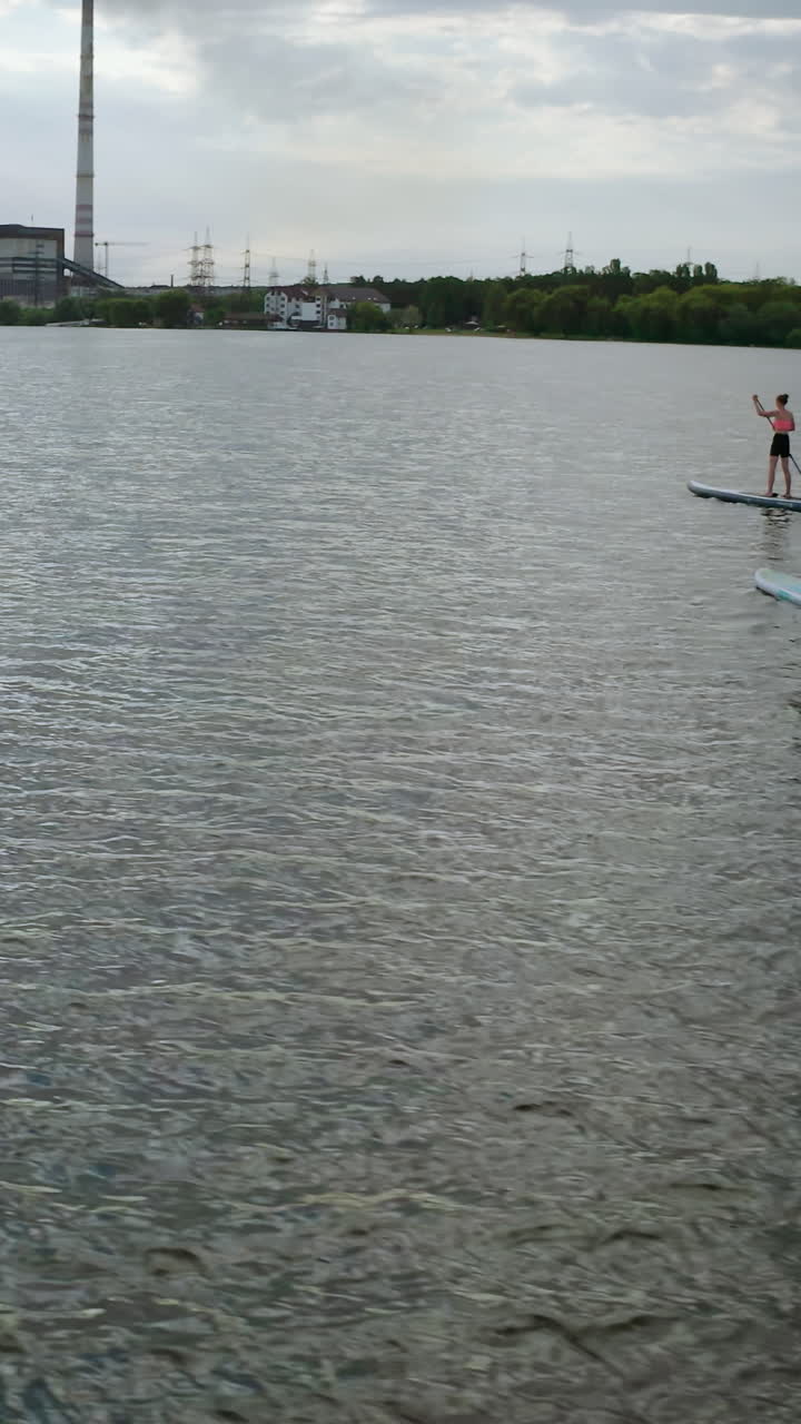 Young people sailing on water. Man and woman travelling on river on paddle boards on industry background in the evening. Summer rest. Vertical video