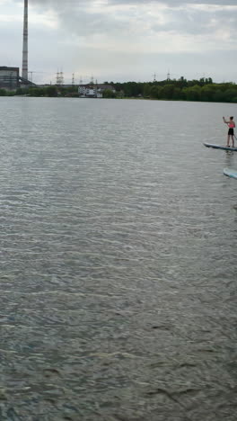 young people sailing on water. man and woman travelling on river on paddle boards on industry background in the evening. summer rest. vertical video