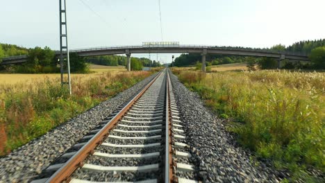 Slow speed aerial drone view at low altitude moving forward following straight railroad tracks with bridge ahead and agricultural fields on both sides, simulating train ride in summer time