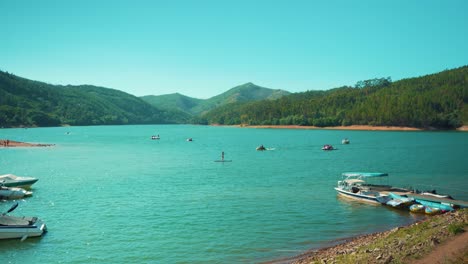 Orilla-Del-Lago-Con-Barcos-Deportivos-De-Muelle-Pasando-Y-Practicando-Paddle-Entre-Colinas-De-Pinos-Al-Sol-4k