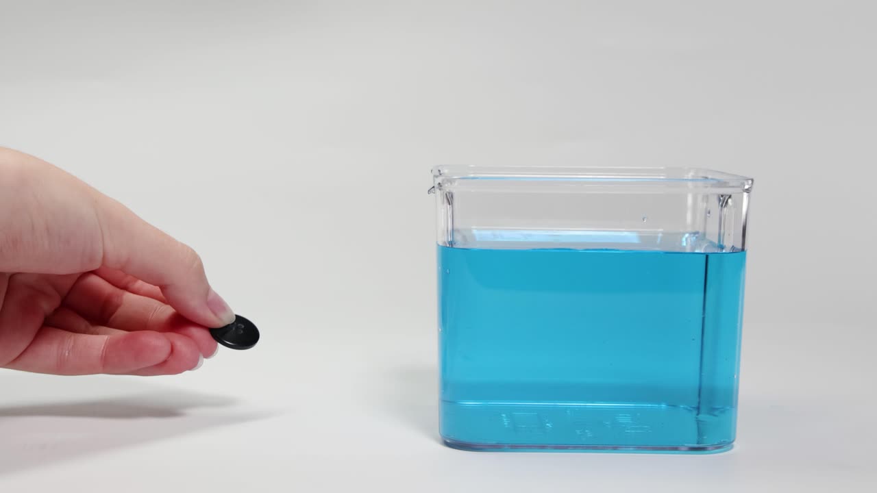 A hand drops a black button into a clear container of blue liquid, demonstrating buoyancy and sinking