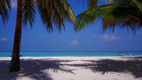 view gets closer to turquoise waters and white beaches of a tropical island as she walks between the two palm trees on a bright day, two small wooden boats have anchored near by