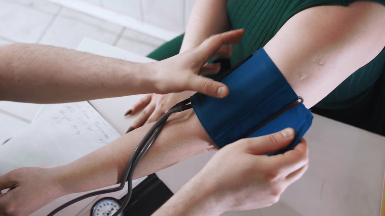 Donna nera seduta al tavolo che riceve un controllo della pressione sanguigna da un infermiere paramedico a domicilio, vestito verde, bracciale applicato al braccio, stetoscopio e manometro visibili, documenti sulla scrivania, cura premurosa.