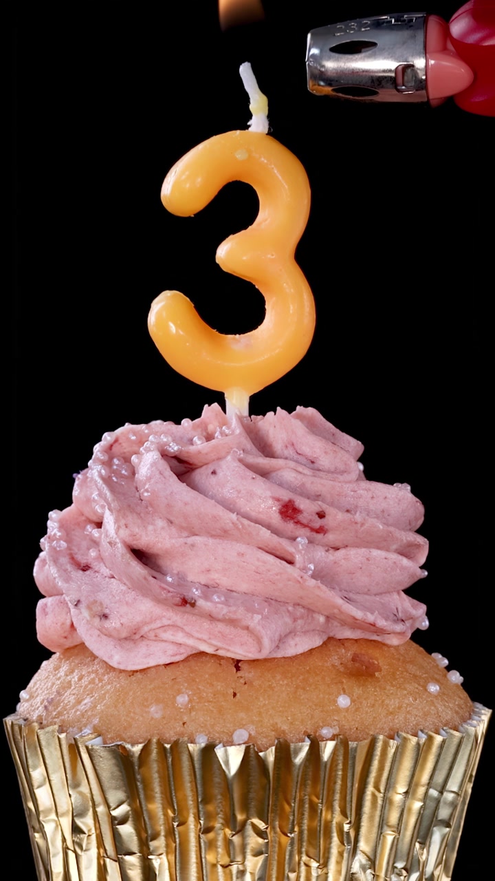 Una mano usa un accendino per accendere una candela di compleanno numero tre sopra un cupcake alla fragola con glassa rosa, su uno sfondo nero con illuminazione drammatica