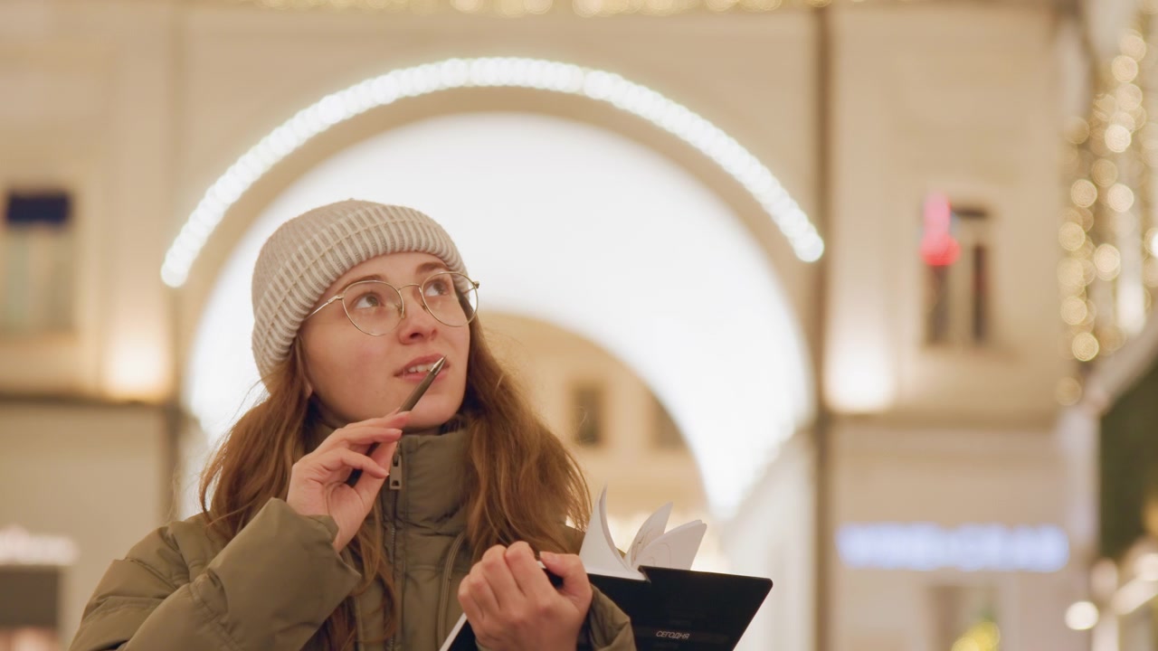 White woman in winter hat musing, holding pen near lips and flipping notebook pages beneath glittering arch, soft smile and distant gaze suggest quiet inspiration and editorial storytelling mood